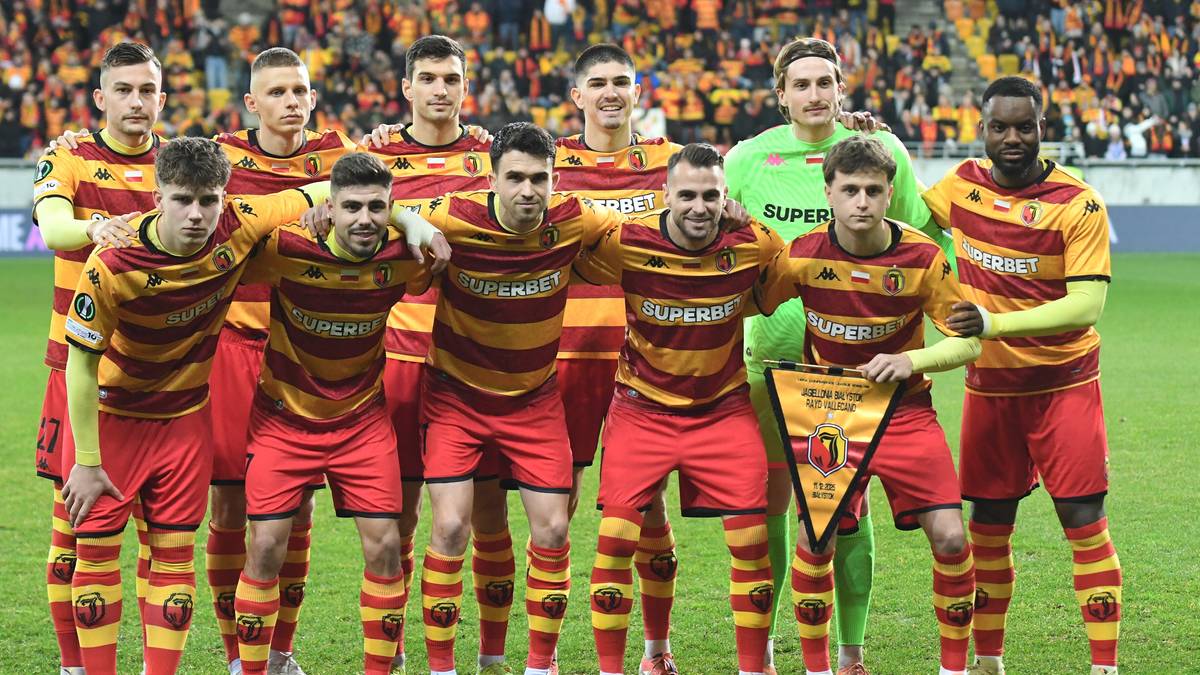 Drużyna piłkarska Jagiellonii Białystok w komplecie, stojąca na murawie stadionu, ubrana w czerwono-żółte stroje. Jeden z zawodników trzyma proporzec.