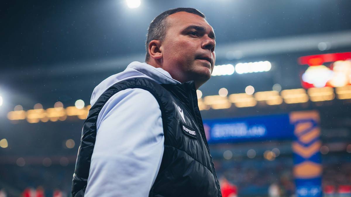 Portret mężczyzny w czarnej kamizelce nałożonej na białą bluzę, stojącego na tle stadionu piłkarskiego. Mężczyzna patrzy w bok z wyrazem koncentracji na twarzy.