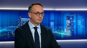 Dariusz Klimczak w garniturze i w okularach na tle niebieskiego ekranu w studio telewizyjnym.