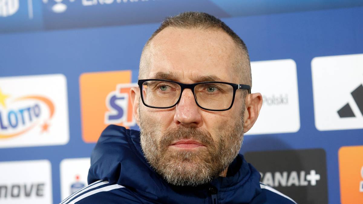 Portret mężczyzny w okularach i kurtce, na tle niebieskiej ściany z logotypami.