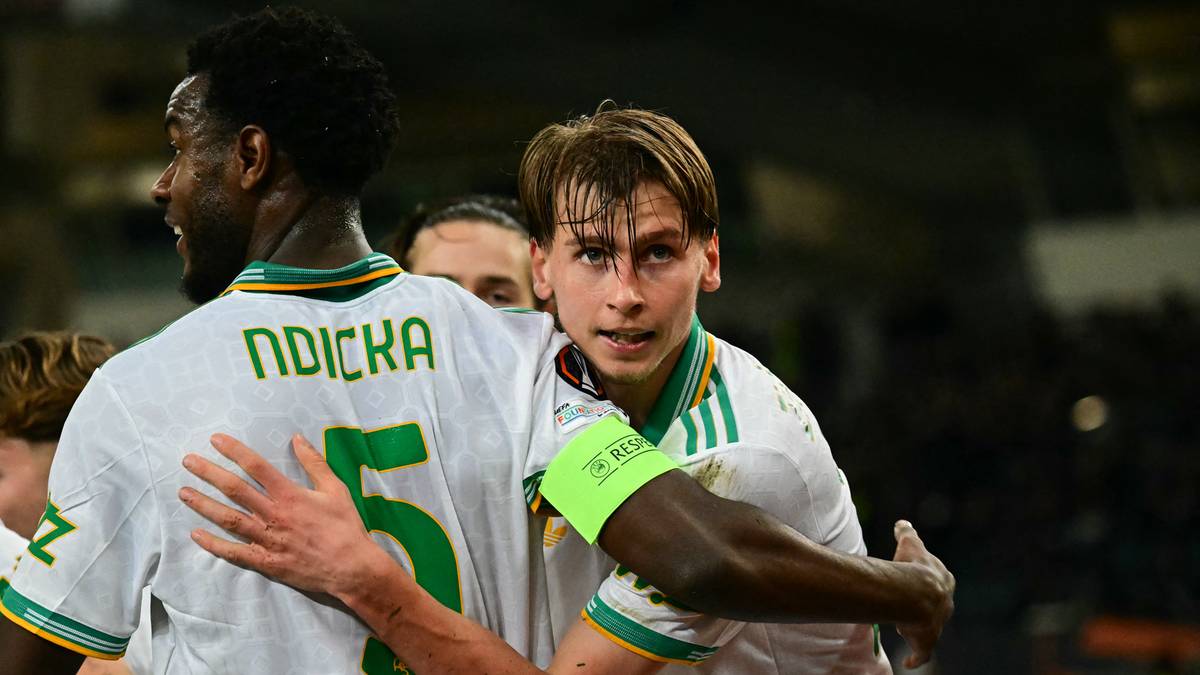 Dw&oacute;ch piłkarzy w białych koszulkach z zielonymi detalami, jeden z nich ma opaskę kapitańską na ramieniu.