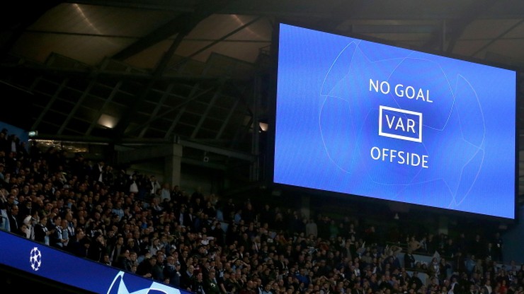 Duży ekran na stadionie piłkarskim pokazuje napis "NO GOAL" (Nie ma gola) oraz "VAR OFFISDE" (VAR spalony). Widoczna jest część trybun wypełnionych kibicami.