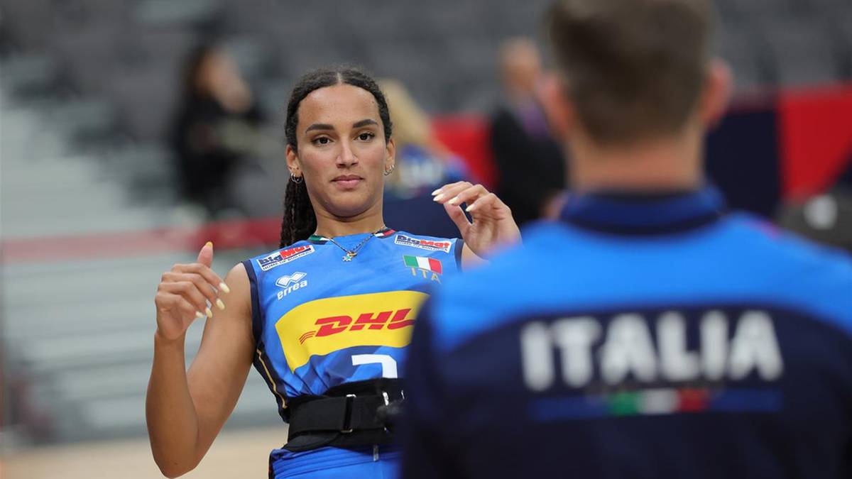 Młoda kobieta w niebiesko-żółtej koszulce z logo DHL i włoską flagą, z uniesionymi kciukami, patrzy w stronę obiektywu. W tle widać zamazaną postać w niebieskiej koszulce z napisem "ITALIA".