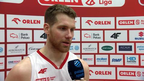 Szymon Tomczak, koszykarz w białej koszulce, udziela wywiadu na tle ściany z logo sponsor&oacute;w.