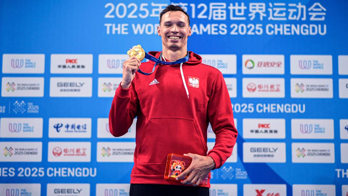 Uśmiechnięty polski sportowiec w czerwonej bluzie z kapturem, trzymający złoty medal i czerwone pudełko, na tle niebieskiej ściany z logo "THE WORLD GAMES 2025 CHENGDU".