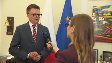 Mężczyzna w garniturze i okularach m&oacute;wi do mikrofonu trzymanego przez kobietę. Za nimi wiszą flagi Polski i Unii Europejskiej, a na ścianie wisi obraz.