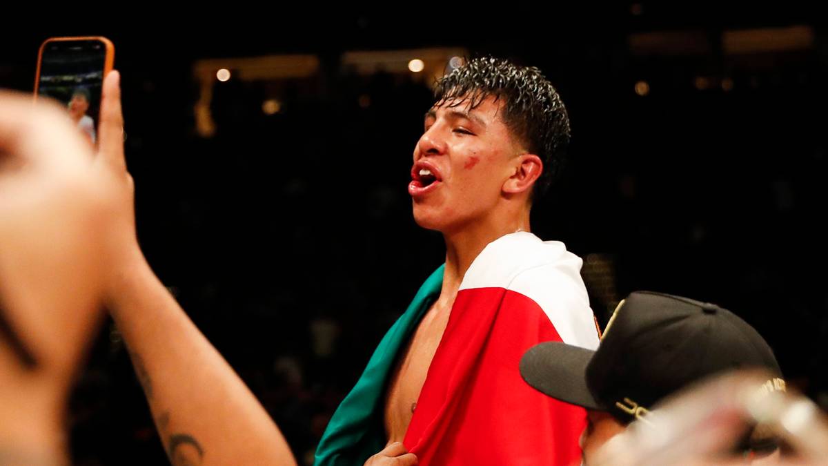 Pięściarz w koszulce z flagą Meksyku podczas walki, ubrany w czerwono-białą szatę, z widoczną kontuzją na twarzy.