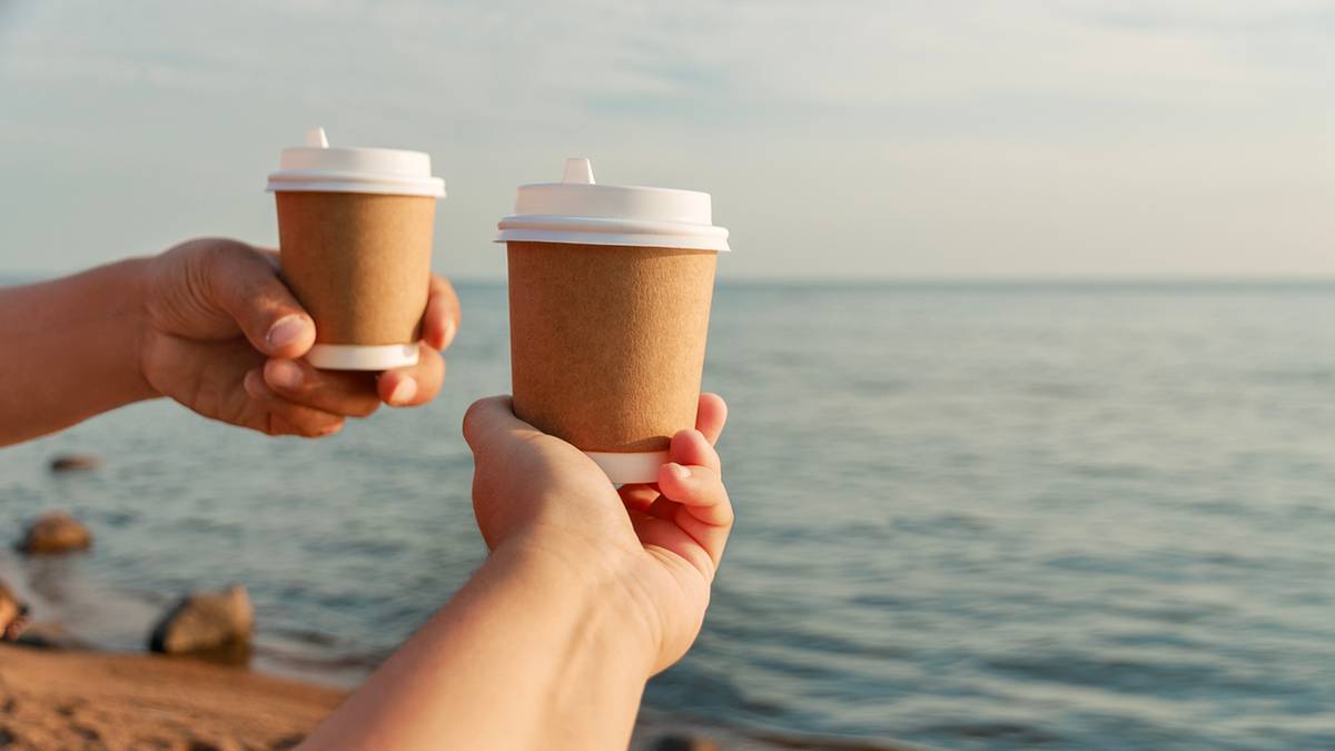 Dwie osoby trzymają w dłoniach papierowe kubki z kawą na tle morza i plaży.