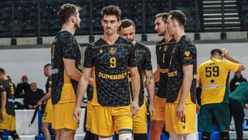 Siatkarze w ż&oacute;łto-czarnych strojach stoją na boisku hali sportowej.