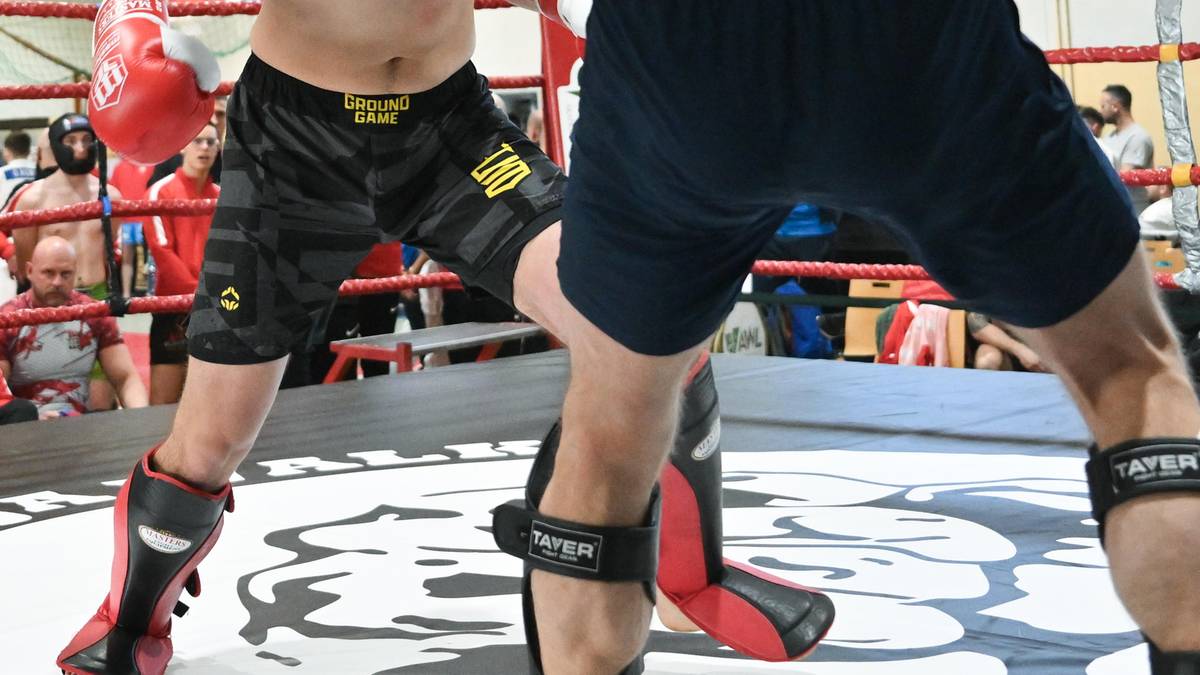 Dw&oacute;ch zawodnik&oacute;w kickboxingu walczy w ringu, jeden z nich atakuje, drugi broni się. W tle sędzia i publiczność.