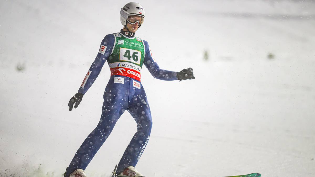 Skoczek narciarski w niebieskim kombinezonie z numerem 46 na piersi stoi na nartach na zaśnieżonej skoczni.