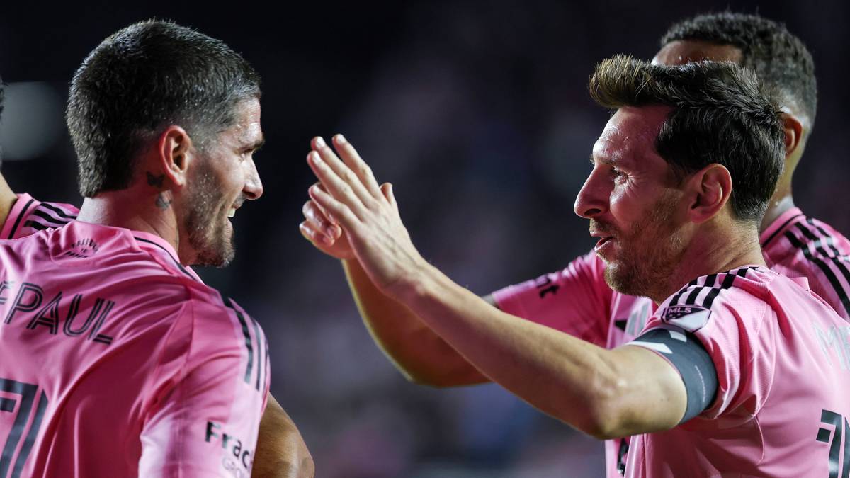 Dwóch piłkarzy w różowych koszulkach świętuje bramkę, jeden z nich, Lionel Messi, podnosi dłoń w geście radości.