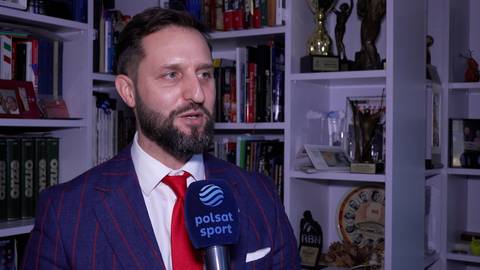 Mężczyzna z brodą w garniturze i czerwonym krawatem, trzymający mikrofon z logo Polsat Sport. W tle p&oacute;łki z książkami i trofeami.