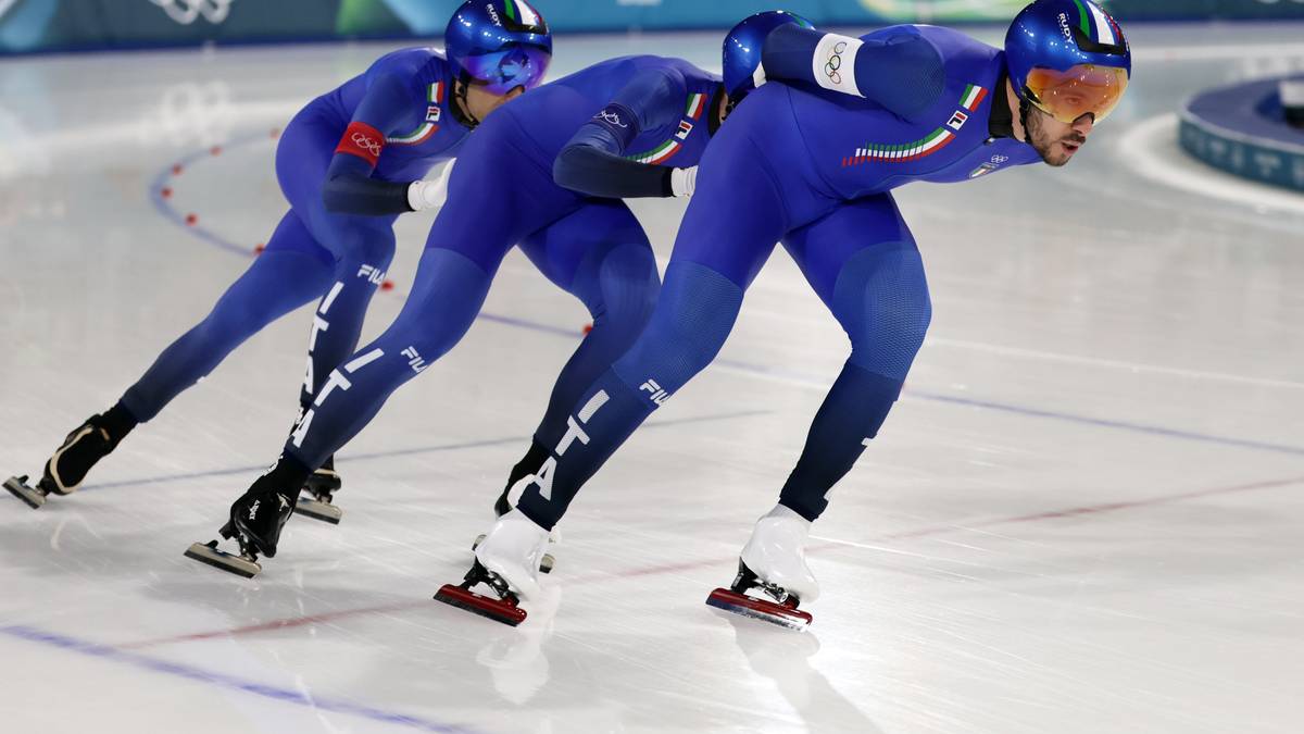 Trzech zawodników w niebieskich strojach i kaskach jedzie na łyżwach po lodowisku w wyścigu.