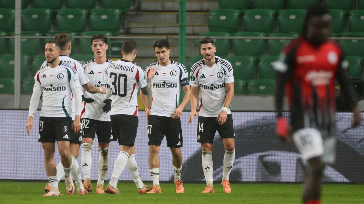 Piłkarze Legii Warszawa w białych strojach świętują bramkę na tle zielonych trybun.