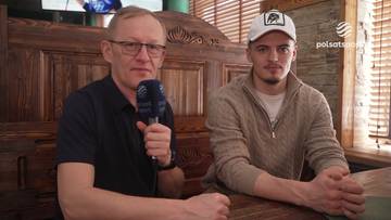 Dwóch mężczyzn siedzi przy drewnianym stole. Jeden z nich, starszy, w okularach, trzyma mikrofon. Drugi, młodszy, w czapce, jest obiektem wywiadu. W tle widoczne logo Polsat Sport.