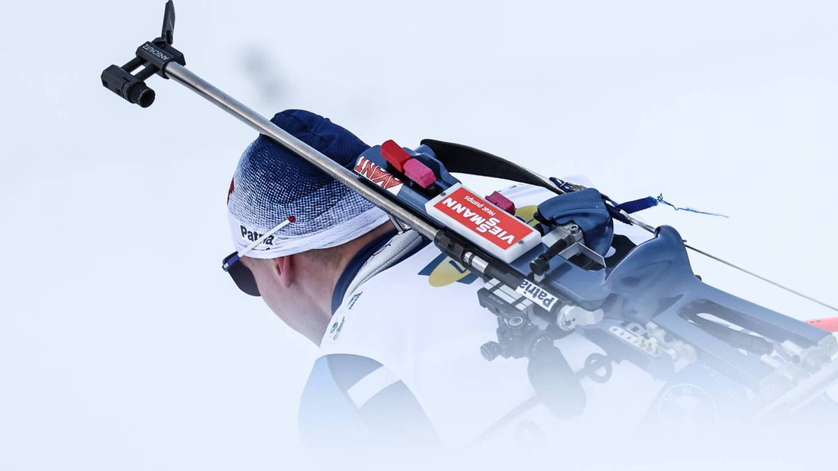 Widok zza pleców biathlonisty podczas zawodów, z karabinem narciarskim na ramieniu. Zawodnik ubrany jest w biały kombinezon, czapkę i gogle.