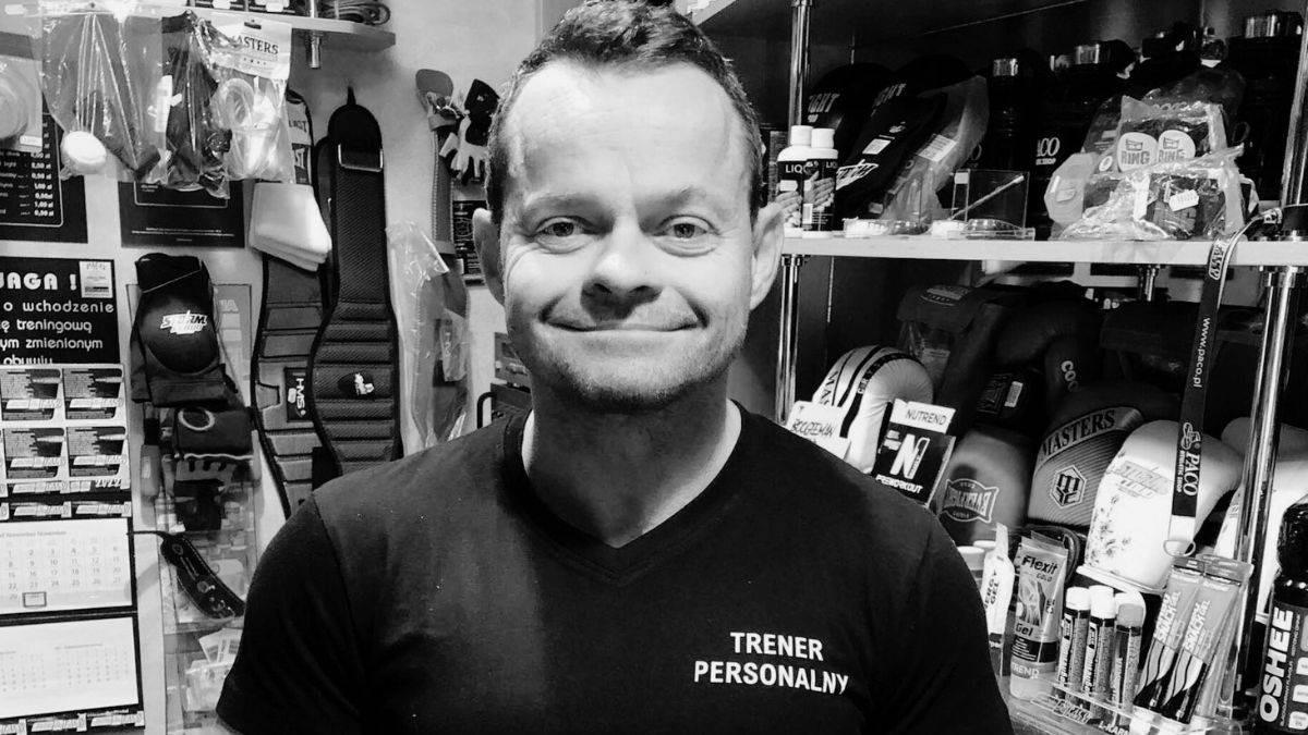 Czarno-białe zdjęcie przedstawiające uśmiechniętego mężczyznę z krótkimi włosami, ubranego w czarną koszulkę z napisem "TRENER PERSONALNY". W tle widoczne są półki sklepowe z różnymi akcesoriami sportowymi.