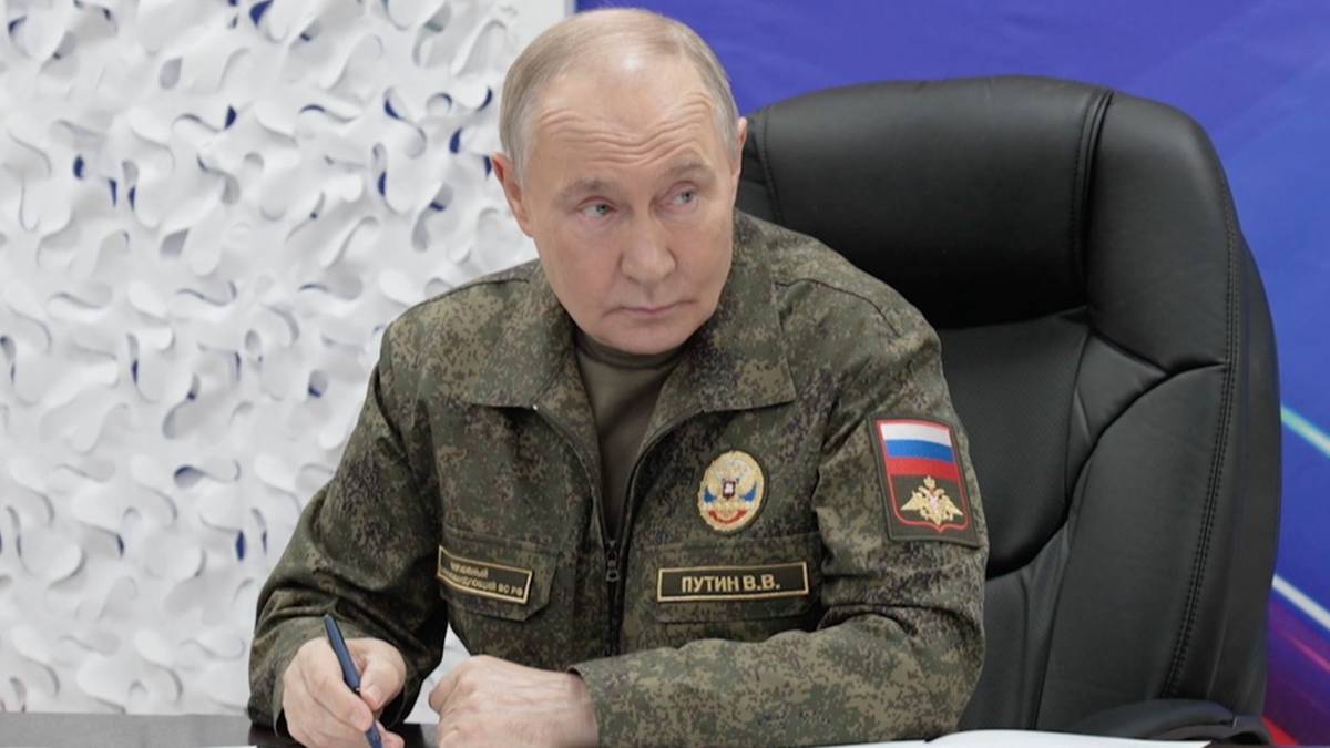 Władimir Putin w mundurze wojskowym siedzi przy biurku i pisze, na niebieskim tle z rosyjskimi napisami.