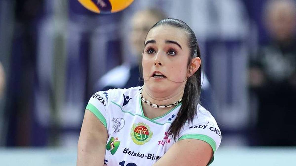 Młoda kobieta, zawodniczka siatk&oacute;wki, w białej koszulce z zielonymi elementami, patrzy w g&oacute;rę, przygotowując się do odbicia piłki.