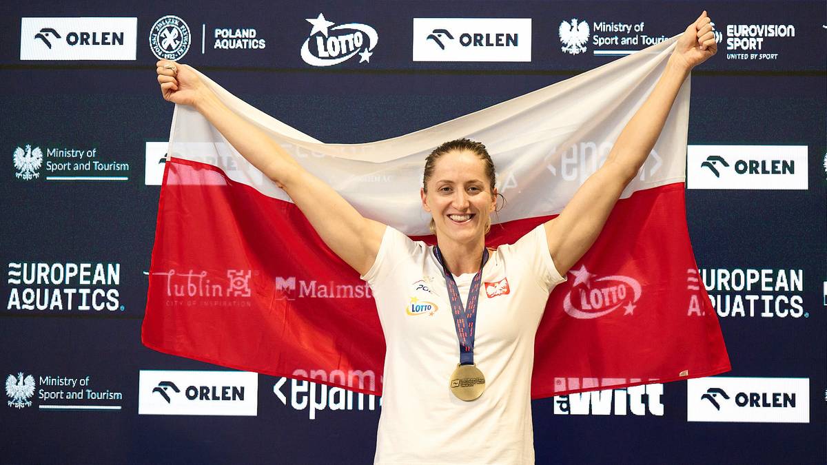 Uśmiechnięta kobieta trzyma rozwiniętą polską flagę za plecami, ubrana jest w białą koszulkę z wpiętym medalem.