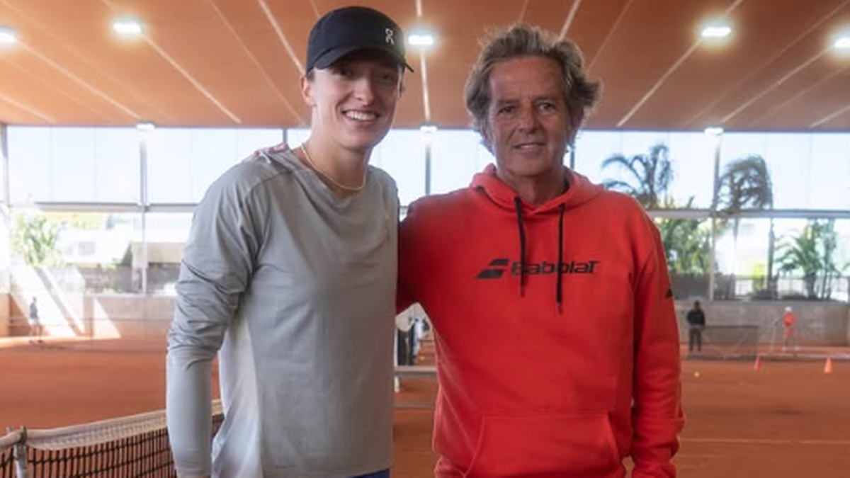 Kobieta i mężczyzna stoją na korcie tenisowym, z uśmiechem na twarzy. Kobieta ubrana jest w szary top i niebieskie spodenki, mężczyzna w pomarańczową bluzę z kapturem i czarne spodnie. Obok nich widoczne są elementy kortu tenisowego i siatka.