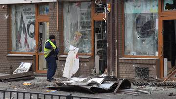 Zniszczony budynek z wybitymi oknami i gruzami na chodniku, w pobliżu stoi policjant w kamizelce odblaskowej.