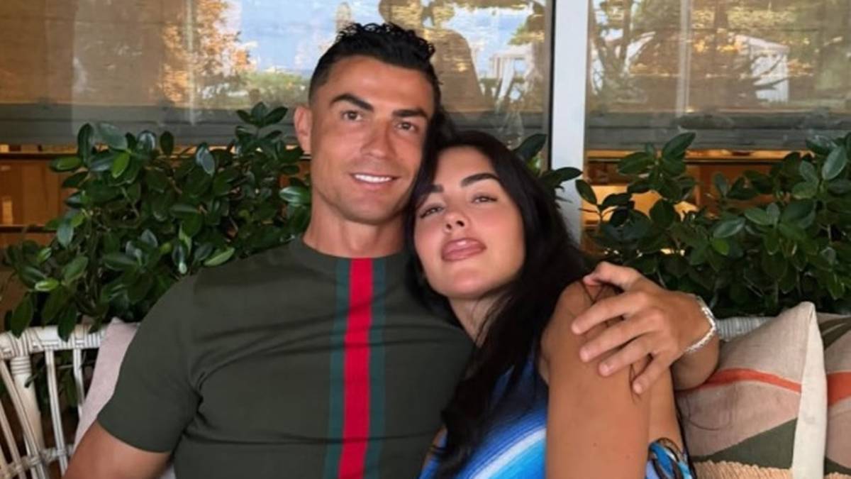 Cristiano Ronaldo i Georgina Rodriguez siedzą blisko siebie, uśmiechnięci. Cristiano ma na sobie zieloną koszulkę z pionowym czerwono-zielonym pasem. Georgina opiera głowę na jego ramieniu, a jej język jest lekko wysunięty.