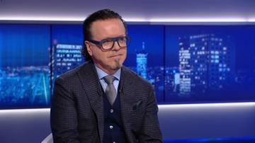 Mężczyzna w szarym garniturze i niebieskich okularach siedzi na kanapie. W tle znajdują się ekrany z widokiem na nocne miasto.