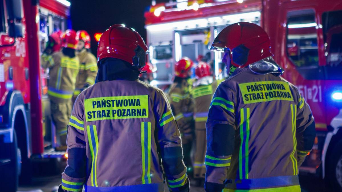 Dw&oacute;ch strażak&oacute;w w odblaskowych kurtkach z napisem "Państwowa Straż Pożarna" i kaskach, stojący na tle woz&oacute;w strażackich.