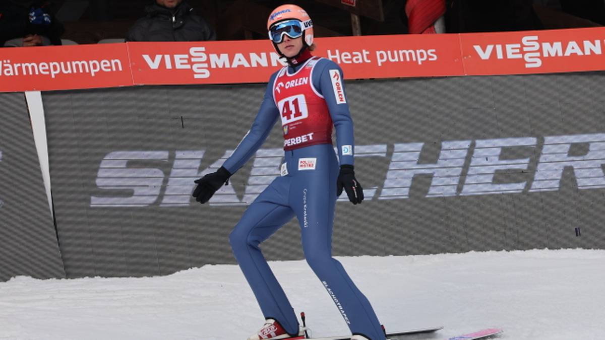 Skoczek narciarski w niebieskim kombinezonie i z numerem 41 na piersi stoi na nartach na śniegu, z rękawicami na dłoniach i goglami na twarzy.