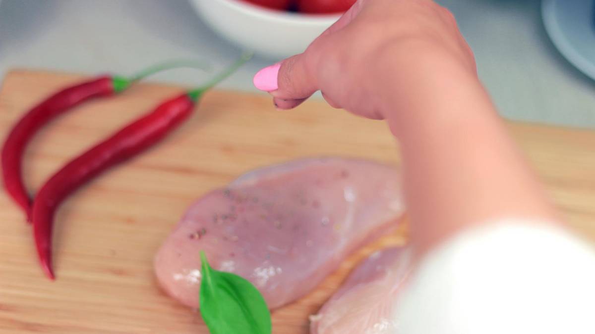 Ręka doprawia surowe piersi kurczaka na desce z chili i ziołami.