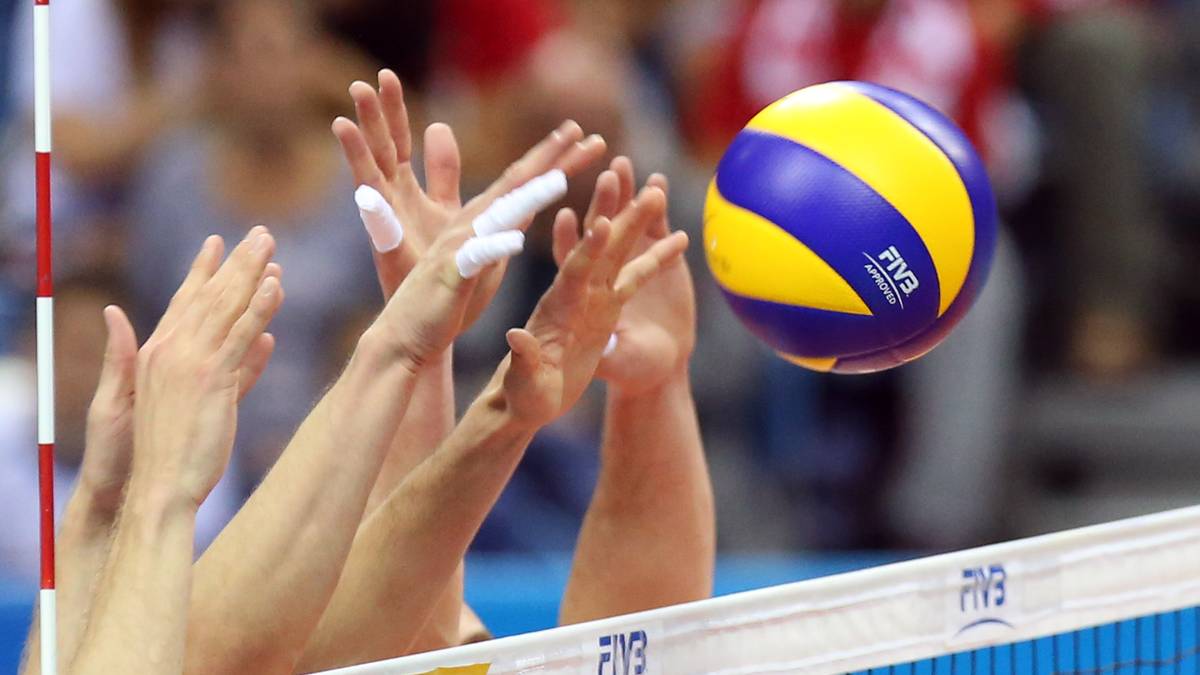 Siatkarze blokują piłkę nad siatką. Piłka w kolorze żółto-niebieskim znajduje się w powietrzu. Widoczni są gracze z podniesionymi rękami.