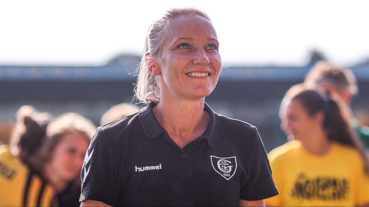 Portret uśmiechniętej kobiety w czarnej koszulce sportowej z herbem drużyny. W tle rozmyte sylwetki innych osób.