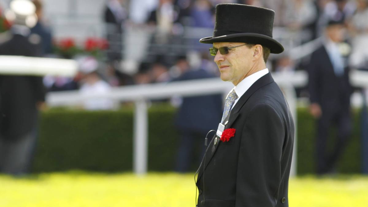 Mężczyzna w cylindrze, okularach przeciwsłonecznych i garniturze patrzy w bok. W tle widać niewyraźną grupę ludzi i białą barierkę.