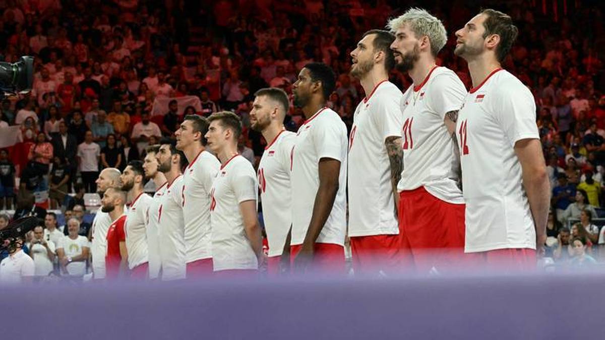 Grupa polskich siatkarzy w białych koszulkach z czerwonymi detalami stoi w rzędzie i słucha hymnu narodowego przed meczem, w tle tłum widzów na trybunach.