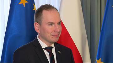 Portret mężczyzny w garniturze i krawacie, stojącego przed polską flagą i flagą Unii Europejskiej.