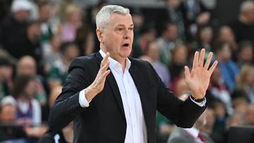 Trener w czarnym garniturze i białej koszuli, z siwymi włosami, gestykuluje rękami podczas meczu koszykówki. W tle widać publiczność.