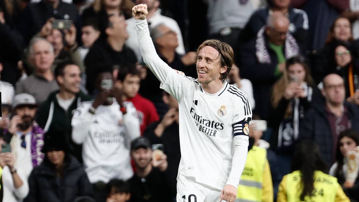 Piłkarz w białej koszulce Realu Madryt z uniesioną ręką świętuje zdobycie bramki, na tle publiczności na stadionie.