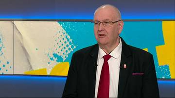 Bogdan Święczkowski w garniturze i czerwonym krawacie podczas wypowiedzi w studio telewizyjnym.
