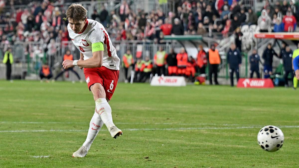 Piłkarz reprezentacji Polski U-21 wykonuje strzał do piłki na zielonym boisku, w tle trybuny z kibicami.