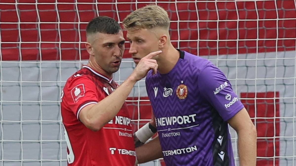 Dwóch piłkarzy w akcji podczas meczu, jeden zawodnik w czerwonej koszulce wskazuje palcem w kierunku zawodnika w fioletowej koszulce.