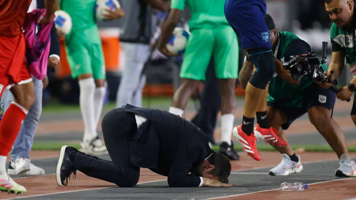 Mężczyzna w garniturze klęczący na bieżni lekkoatletycznej wśród piłkarzy.