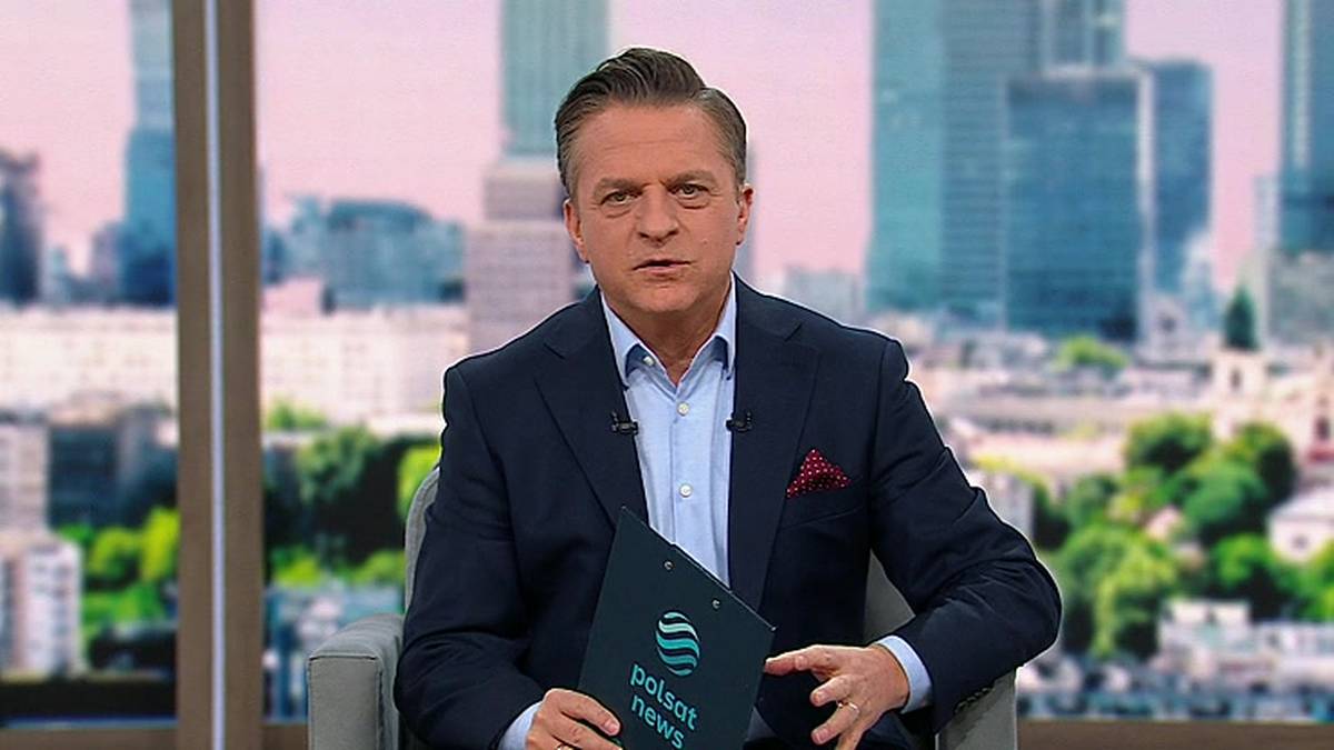Mężczyzna w garniturze siedzi w studiu telewizyjnym, trzymając w rękach teczkę z logo "Polsat News". W tle widoczne są wieżowce i niebo.