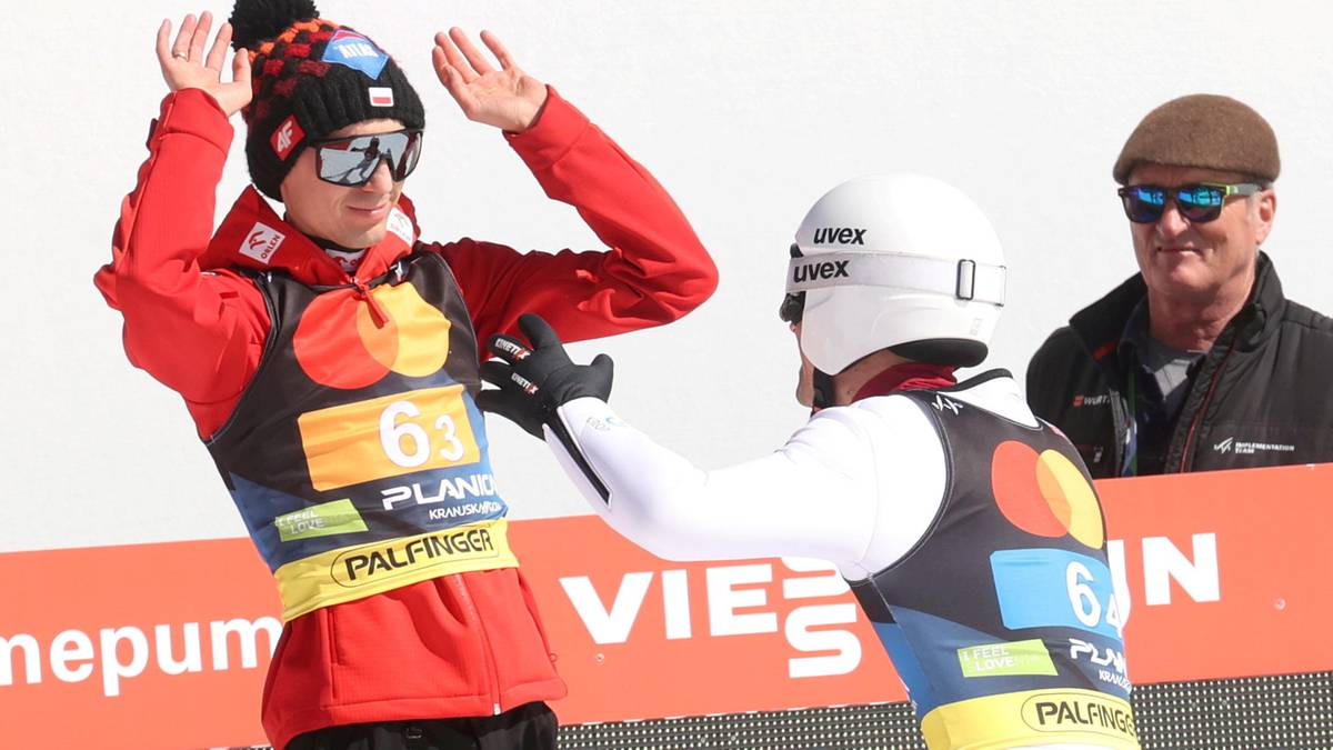 Dwóch skoczków narciarskich w strojach sportowych, jeden z nich w czerwonej kurtce i czapce z pomponem, drugi w białym kombinezonie, pozdrawiają się na podium.