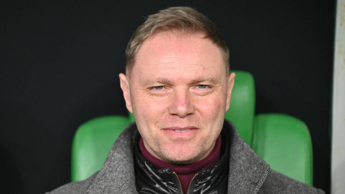 Uśmiechnięty mężczyzna w średnim wieku, ubrany w szary płaszcz i ciemną kurtkę, siedzi na tle zielonych siedzeń.