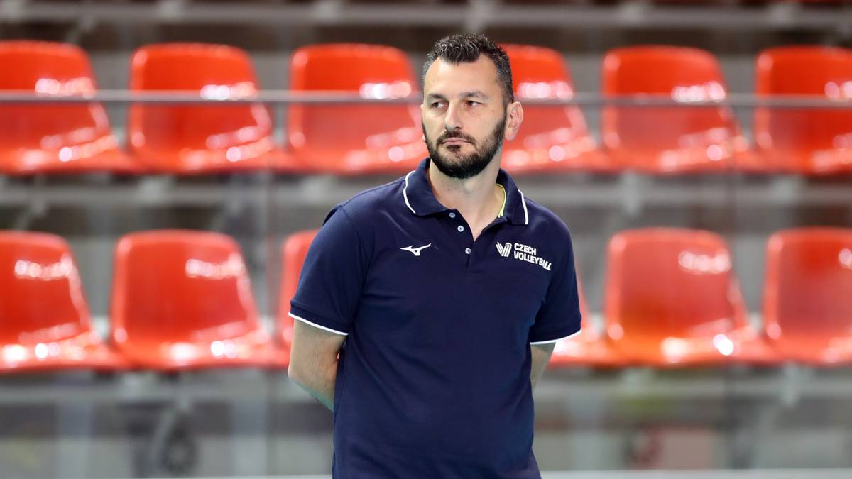 Mężczyzna w granatowej koszulce polo z kołnierzykiem stoi przed rzędem czerwonych krzesełek na trybunach hali sportowej.