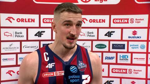 Koszykarz w czerwono-niebieskim stroju sportowym udziela wywiadu przed ścianką sponsorów.