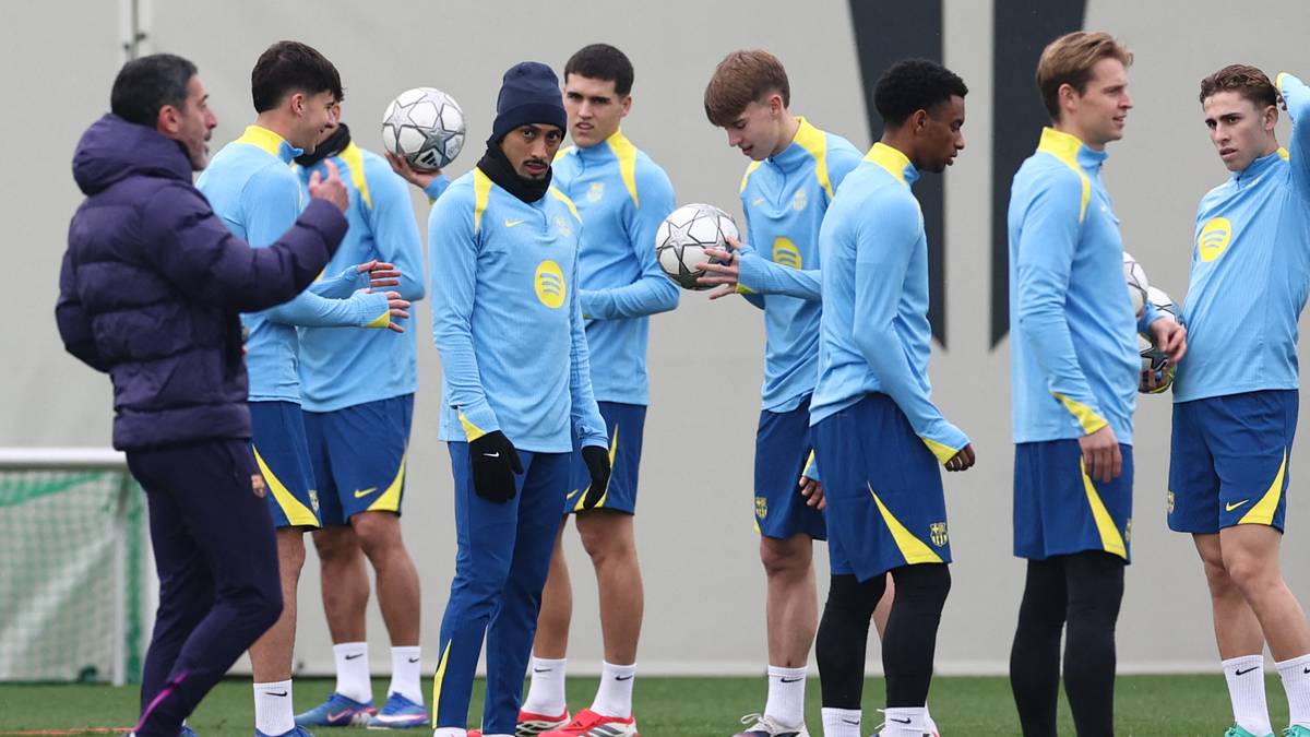 Grupa piłkarzy w niebieskich strojach treningowych zebrana wok&oacute;ł trenera na boisku piłkarskim.