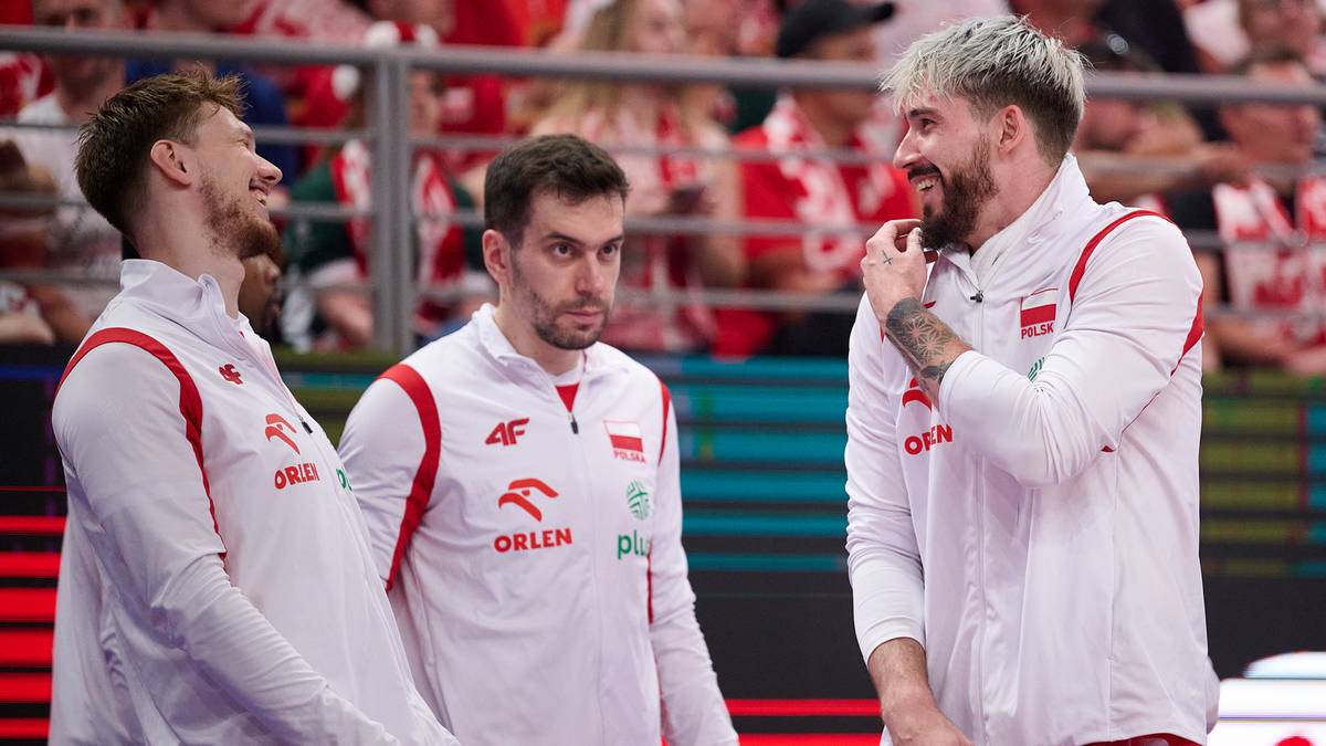 Trzech siatkarzy w biało-czerwonych strojach z logo "ORLEN" na piersiach stoi blisko siebie, rozmawiając. Dwóch z nich jest zwróconych do siebie, jeden lekko się uśmiecha, poprawiając sobie włosy. Tło stanowi trybuna z widzami.